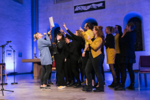 Ein Chor in schwarz-gelber Kleidung zeigt begeistert auf die Urkunde vom Landeschorwettbewerb Bremen, alle freuen sich.