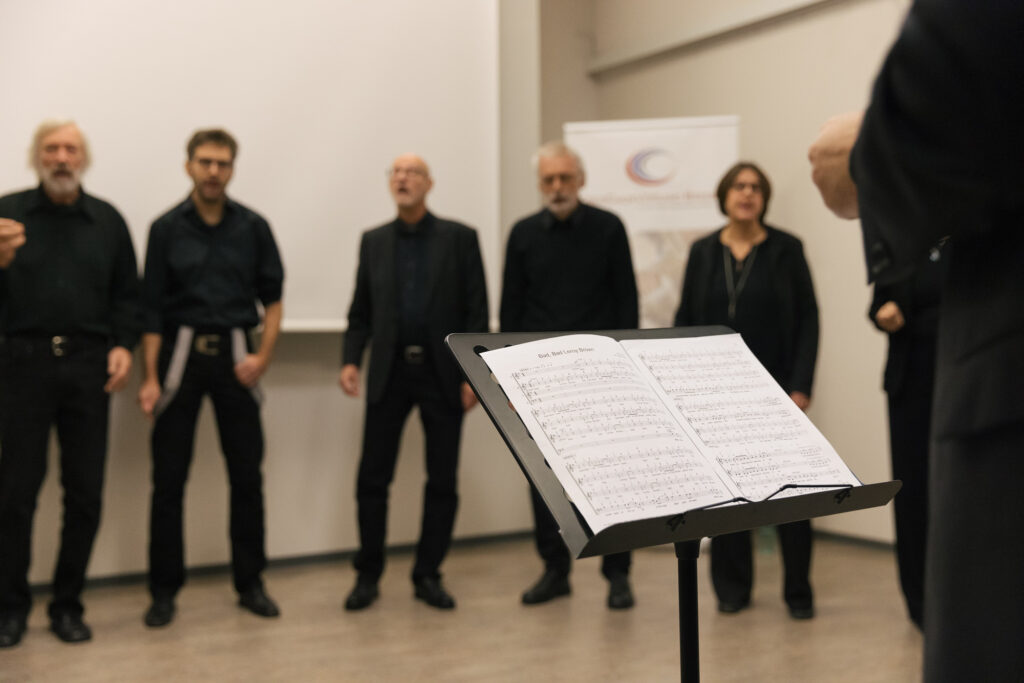 Blick auf einen Notenständer mit Chornoten, dahinter stehen fünf Menschen nebeneinander und singen