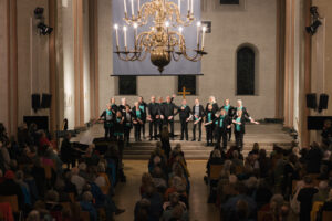 Ein Chor in schwarz-grüner Kleidung steht singend und mit ausgebreiteten Armen auf den Altarstufen der Kulturkirche St. Stephani