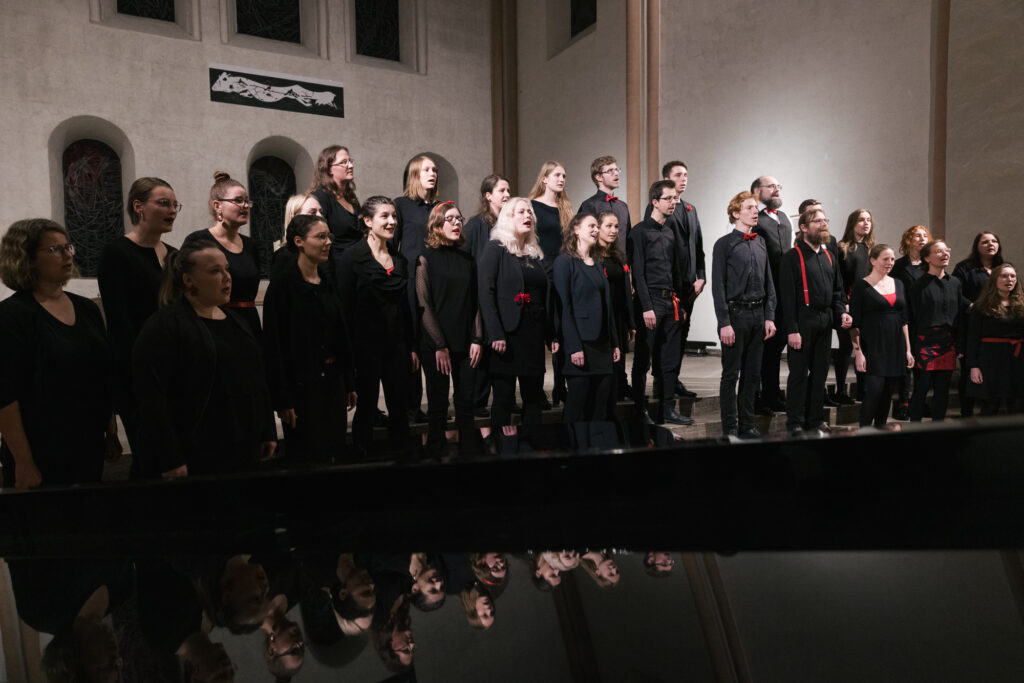 Ein Chor in schwarzer Kleinung und roten Schmuck-Elementen steht auf den Altarstufen der Kulturkirche St. Stephani, Gesichter spiegeln sich im Flügeel