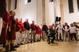 Ein Chor in rot-weiß gekleidet steht im Halbkreis und singt mit ausgebreiteten Armen, ein Akkordeonist sitzt auf einem Stuhl vor dem Chor