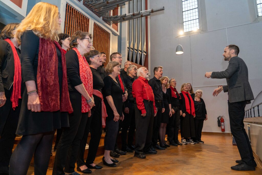 Ein rot-schwarz gekleideter gemischter Chor steht singend auf der Orgelempore der Kulturkirche St. Stephani und wird von einem Mann in grau-schwarzer Kleidung dirigiert