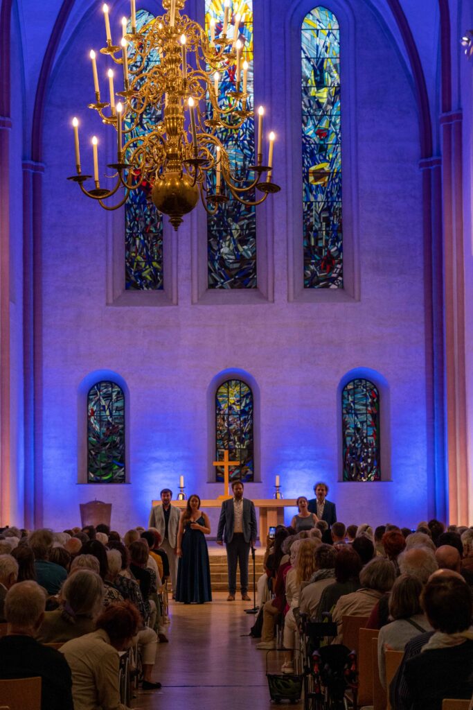 Zwei Frauen und drei Männer stehen nebeneinander singend vor dem Hochaltar in der Kulturkirche St. Stephani, Blick von hinten in den Kirchenraum, einschließlich bunte Kirchenfenster im Hintergrund und Deckenleuchter