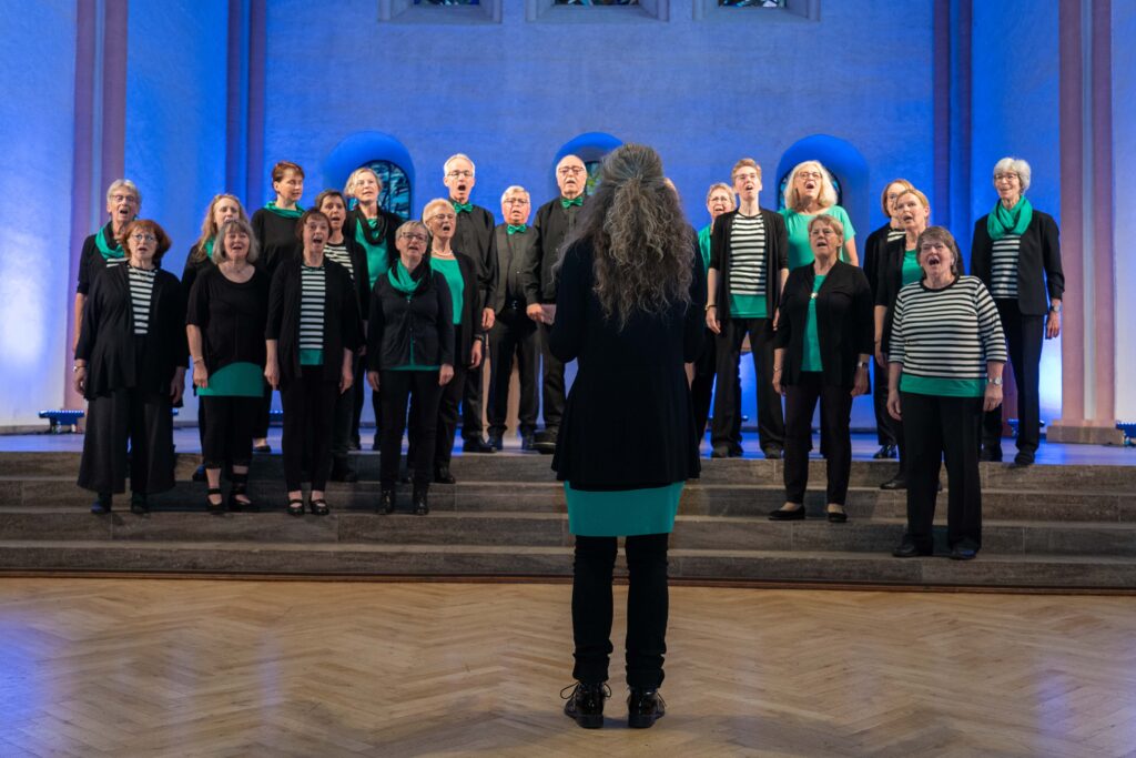 Ein gemischter Chor steht singend auf den Stufen des Hochaltars in der Kulturkirche St. Stephani. Die Frauen tragen schwarz-weiß gestreifte oder grüne Shirts und haben grüne Schals um, die Männer tragen grüne Fliegen. Die Chorleiterin steht mit dem Rücken zum Publikum.
