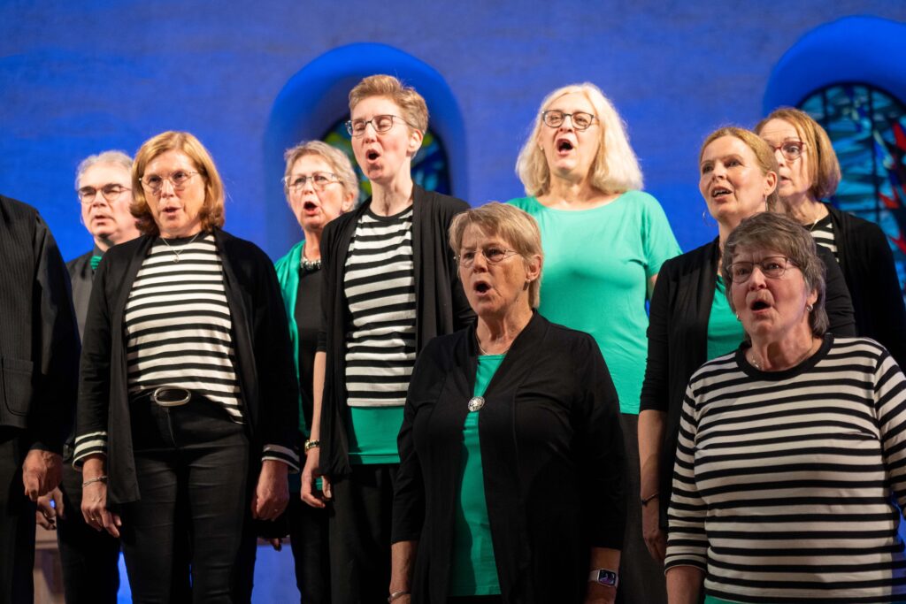 Nahaufnahme eines singenden Chors. Die abgebildeten Frauen tragen schwarz-weiß gestreifte oder grüne Shirts.