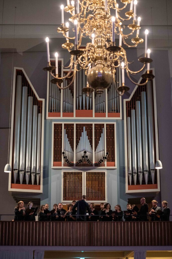 Ein schwarz gekleideter gemischter Kammerchor steht auf der Orgelempore der Kulturkirche St. Stephani, Blick von unten, im Hintergrund eine große Orgel, im Vordergrund ein Deckenleuchter