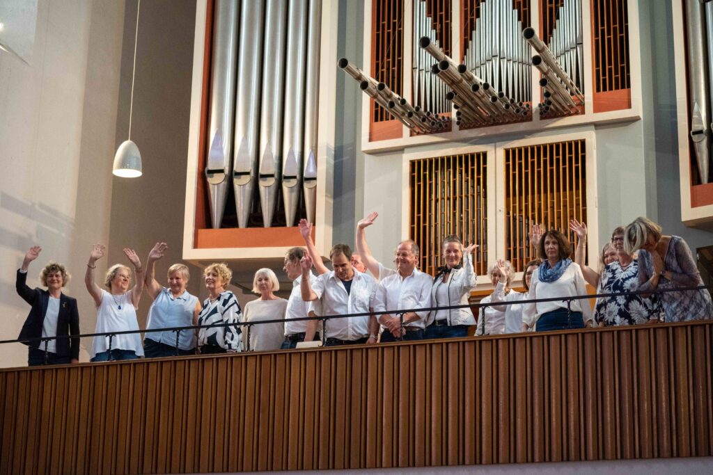 Ein hell gekleideter gemischter Chor steht an der Brüstung der Orgelempore in der Kulturkirche St. Stephani und winkt dem Publikum zu, Blick von unten, im Hintergrund eine große Orgel