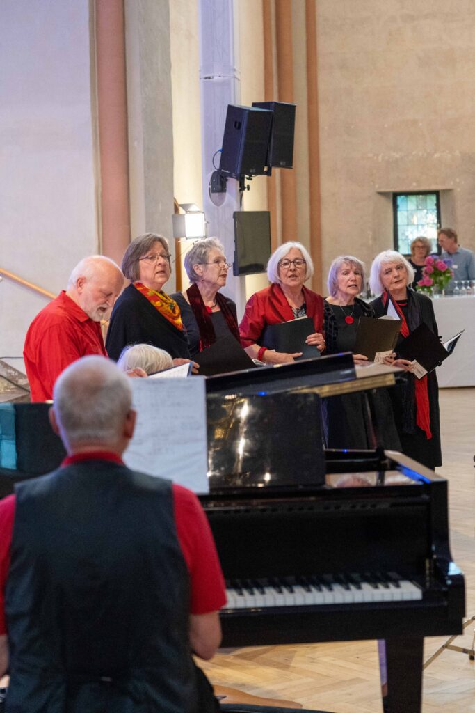 Ein rot-schwarz gekleidete Vokalensemble steht singend in der Kulturkirche St. Stephani, ein Mann sitzt am Flügel und begleitet die Singenden