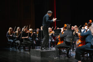 Blick in ein Orchester aus jungen, schwarz gekleideten Menschen, die konzentriert ihre Instrumente spielen. Vor ihnen steht der Dirigent auf einem Podest.