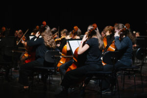 Rückenansicht von schwarz gekleideten, sitzenden Menschen an Violoncelli.