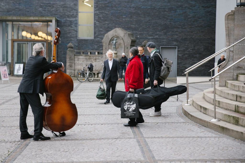 Eine Gruppe von Musikern steht vor dem Eingang des Stadttheaters, die Menschen haben Instrumentenkoffer und Taschen in den Händen. Links lehnt ein Musiker auf seinem Kontrabass und wendet dem Betrachter den Rücken zu.