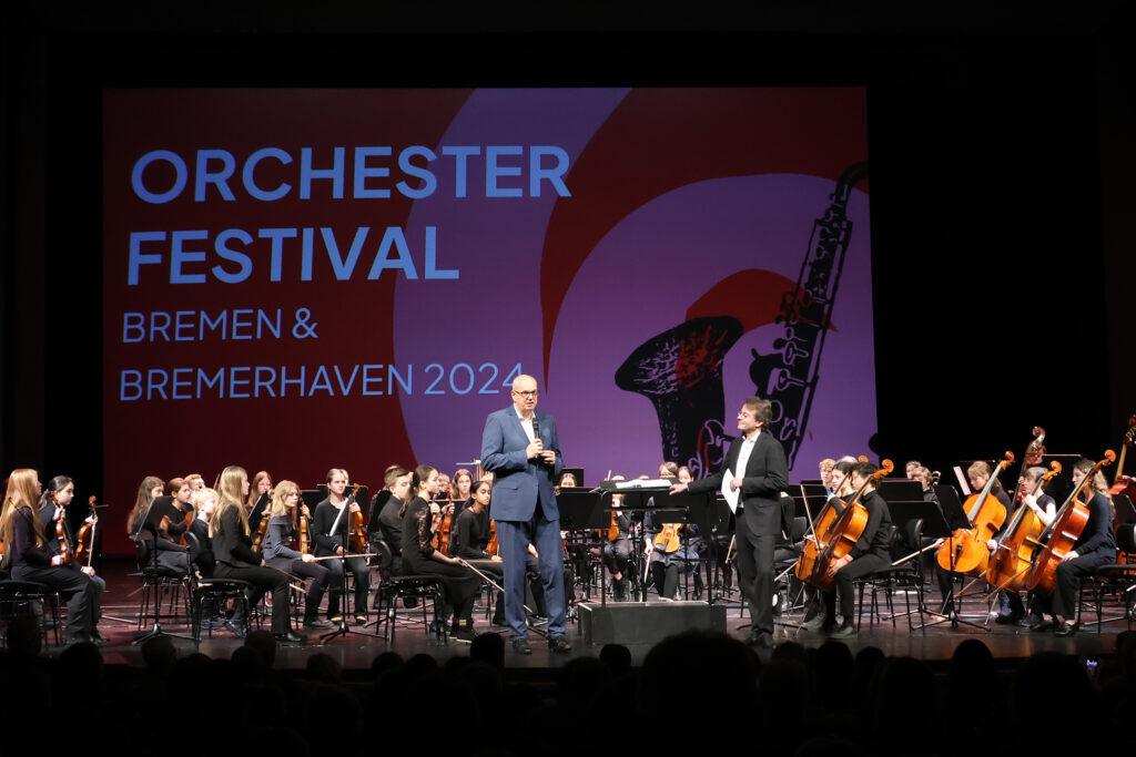 Ein Jugendorchester sitzt auf der Bühne. Vor den Musikerinnen und Musikern steht Bürgermeister Dr. Andreas Bovenschulte und spricht, der Vorsitzende vom Landesmusikrat Marc Niemann hört zu. Im Hintergrund ist der Schriftzug "Orchesterfestvial Bremen & Bremerhaven 2024" zu sehen.