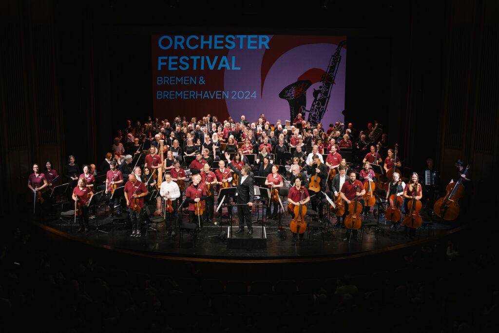 200 Musikerinnen und Musiker stehen mit ihren Instrumenten auf der Bühne des Stadttheaters Bremerhaven, im Hintergrund ist der Schriftzug "Orchesterfestival Bremen & Bremerhaven 2024" zu lesen.