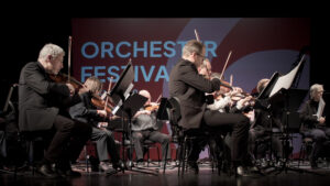 Aufnahme von Orchestermusikern, die konzentriert ihre Streichinstrumente spielen und in die Noten blicken. Im Hintergrund ist der Schriftzug "Orchesterfestival" zu sehen.