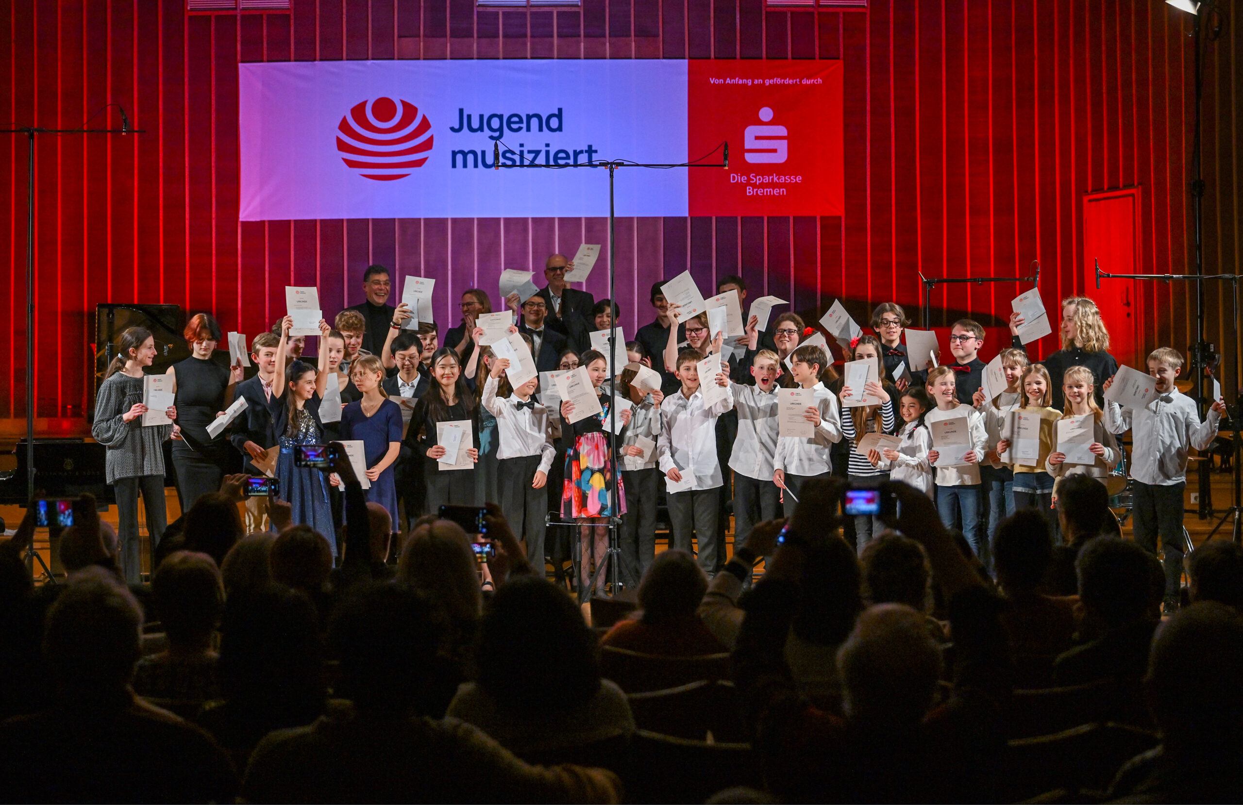 Eine Gruppe Kinder und Jugendlicher stehen zusammen auf der Bühne des Sendesaals Bremen und halten fröhlich ihre Urkunden in die Höhe. Im Hintergrund ist ein Banner mit dem Jugend musiziert-Logo und dem Logo der Sparkasse Bremen zu sehen.