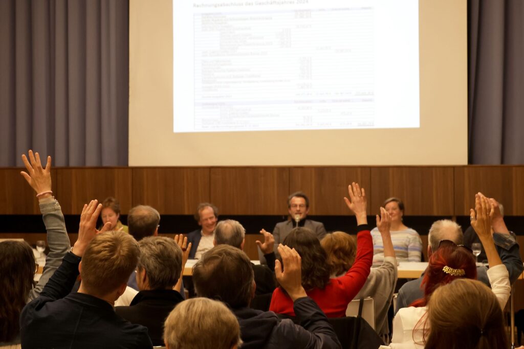 Man sieht viele Menschen von hinten, die in einer Abstimmung die Hand heben, im Hintergrund sind an einem Tisch vier Personen von vorn und an der Wand eine Präsentation zu sehen.
