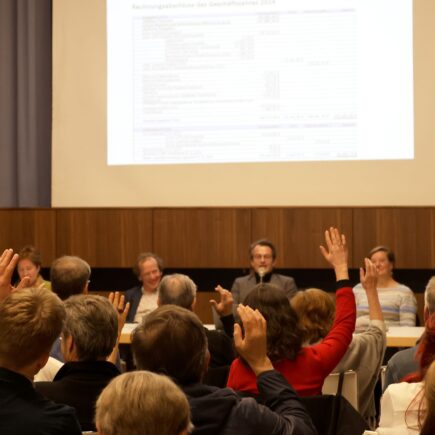 Man sieht viele Menschen von hinten, die in einer Abstimmung die Hand heben, im Hintergrund sind an einem Tisch vier Personen von vorn und an der Wand eine Präsentation zu sehen.