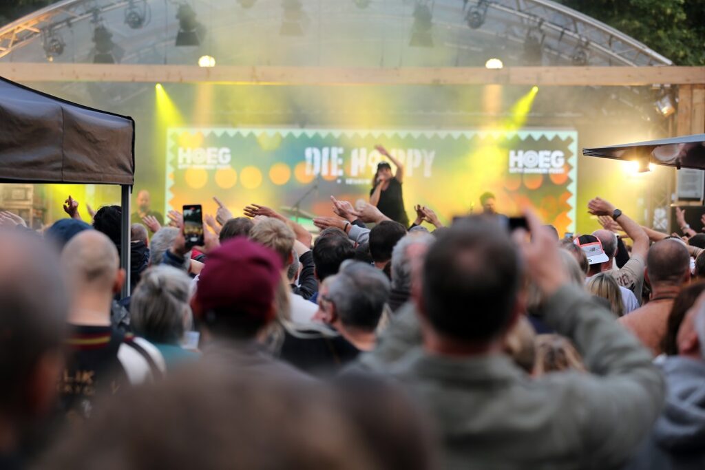 Auf einer Open Air-Bühne ist eine Musikerin zu sehen. Vor der Bühne stehen viele Menschen und hören der Musik zu.