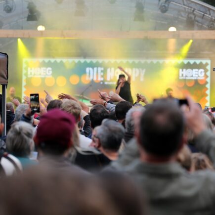 Auf einer Open Air-Bühne ist eine Musikerin zu sehen. Vor der Bühne stehen viele Menschen und hören der Musik zu.
