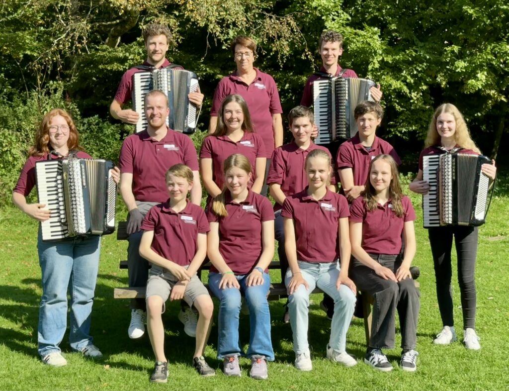 Ein Gruppe Jugendlicher in bordeauxroten T-Shirts schauen lächelnd in die Kamera, vier Personen halten Akkordeons in den Händen