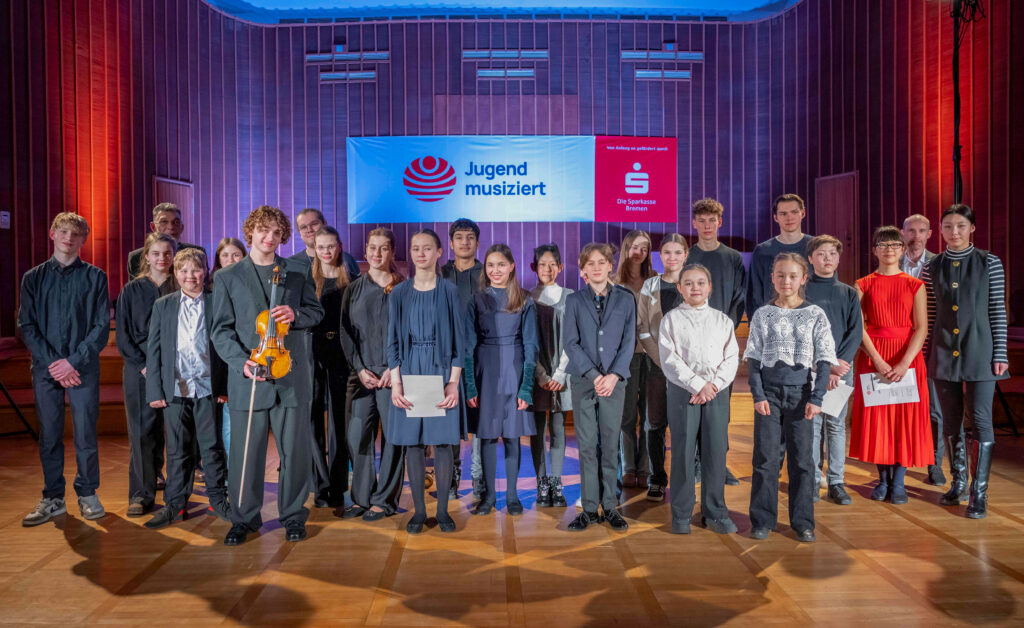 Eine Gruppe Kinder und Jugendlicher steht auf der Bühne des Sendesaals Bremen und blickt fröhlich in die Kamera, im Hintergrund ist ein rot-weißes Banner zu sehen, auf dem Schriftzug und Logo von Jugend musiziert stehen, daneben ist das Logo der Sparkasse Bremen abgebildet.
