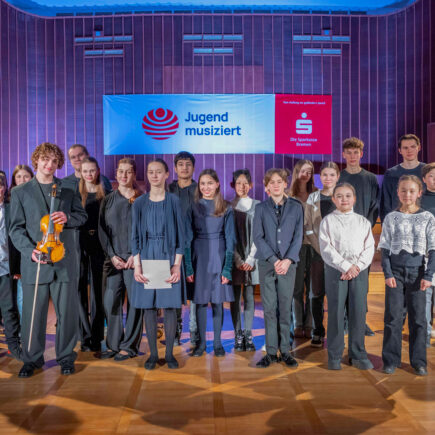 Eine Gruppe Kinder und Jugendlicher steht auf der Bühne des Sendesaals Bremen und blickt fröhlich in die Kamera, im Hintergrund ist ein rot-weißes Banner zu sehen, auf dem Schriftzug und Logo von Jugend musiziert stehen, daneben ist das Logo der Sparkasse Bremen abgebildet.