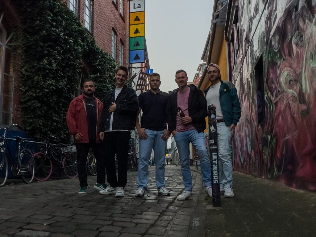 Fünf junge Männer stehen nebeneinander in einer kleinen Straße und schauen in die Kamera, im Hintergrund ist Graffiti an der Wand und ein Banner an einer Hausfassade zu sehen.