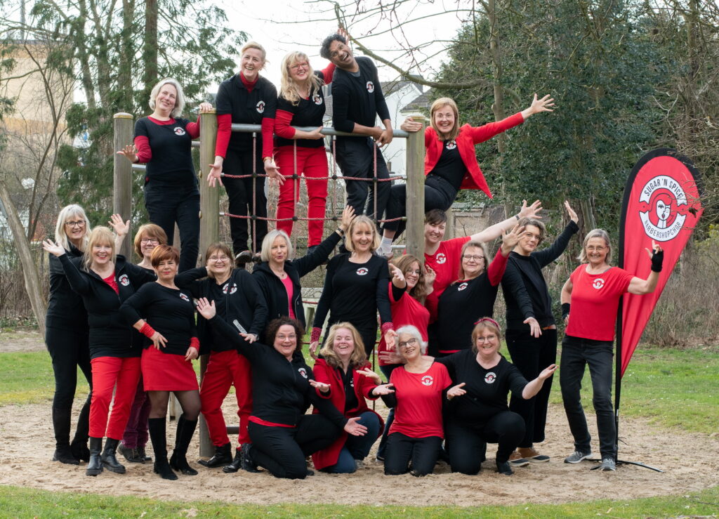 Eine Gruppe schwarz-rot gekleideter Frauen steht an und auf einem Klettergerüst. Sie lachen, winken und strecken ihre Hände aus. Oben auf dem Gerüst ist ein auch ein Mann in schwarzer Kleidung zu sehen. Rechts neben der Gruppe ist ein Banner in den gleichen Farben aufgebaut, darauf steht: Sugar'n'Spice Barbershopchor.