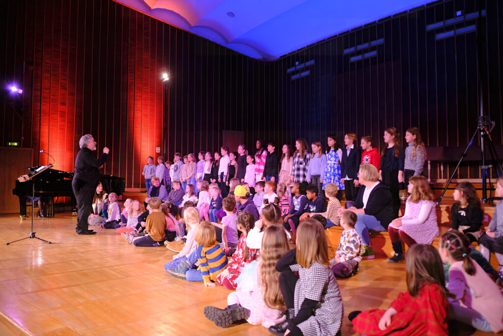 Eine große Gruppe bunt gekleideter Kinder sitzen und stehen auf der Bühne des beleuchteten Sendesaals. Sie schauen den Mann in schwarzer Kleidung an, der vor ihnen steht. Im Hintergrund ist ein schwarzer Flügel zu sehen.