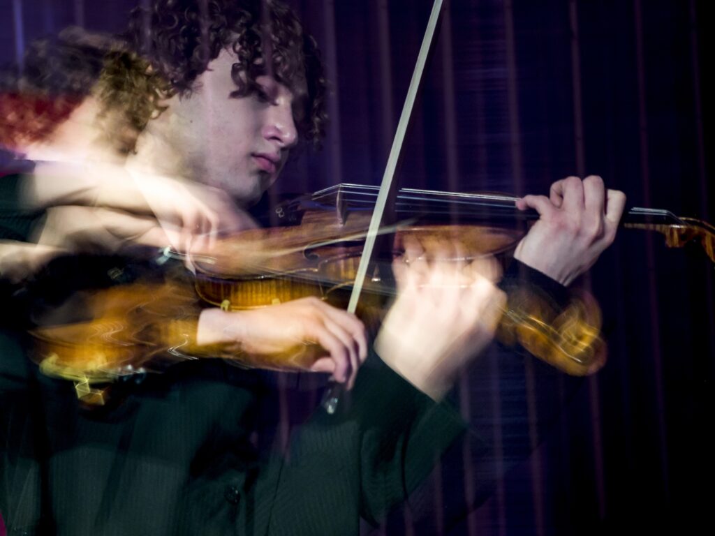 Detailaufnahme eines jungen Mannes mit gelockten braunen Haaren. Er spielt auf einer Violine. Durch eine Fotomontage ist die Person doppelt und teilweise verschwommen zu sehen.