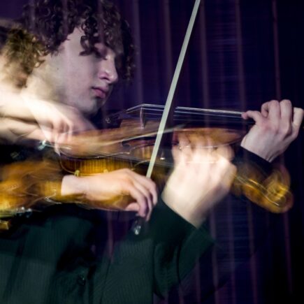Detailaufnahme eines jungen Mannes mit gelockten braunen Haaren. Er spielt auf einer Violine. Durch eine Fotomontage ist die Person doppelt und teilweise verschwommen zu sehen.