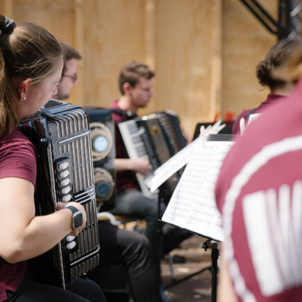 Seitliche Detailaufnahme von Jugendlichen, die Akkordeon spielen, teilweise sind die Menschen unscharf abgebildet.