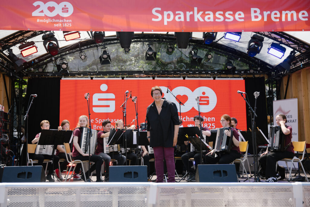 Eine Gruppe Jugendlicher sitzen mit Akkordeons auf einer Open Air Bühne, die Leiterin blickt lachend ins Publikum. Die Bühne ist mit roten Sparkassen-Schriftzügen geschmückt.