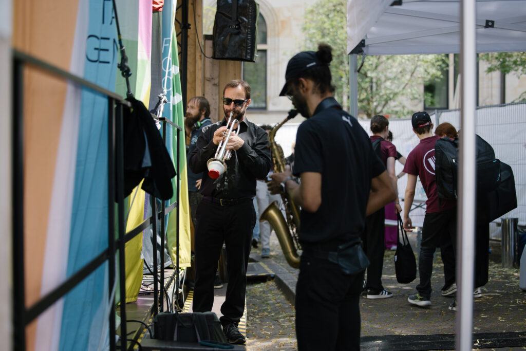 Mehrere Menschen stehen mit ihren Instrumenten hinter der Open Air Bühne, zwei Männer spielen sich an Trompete und Saxofon ein, andere tragen ihre Akkordeons auf dem Rücken.