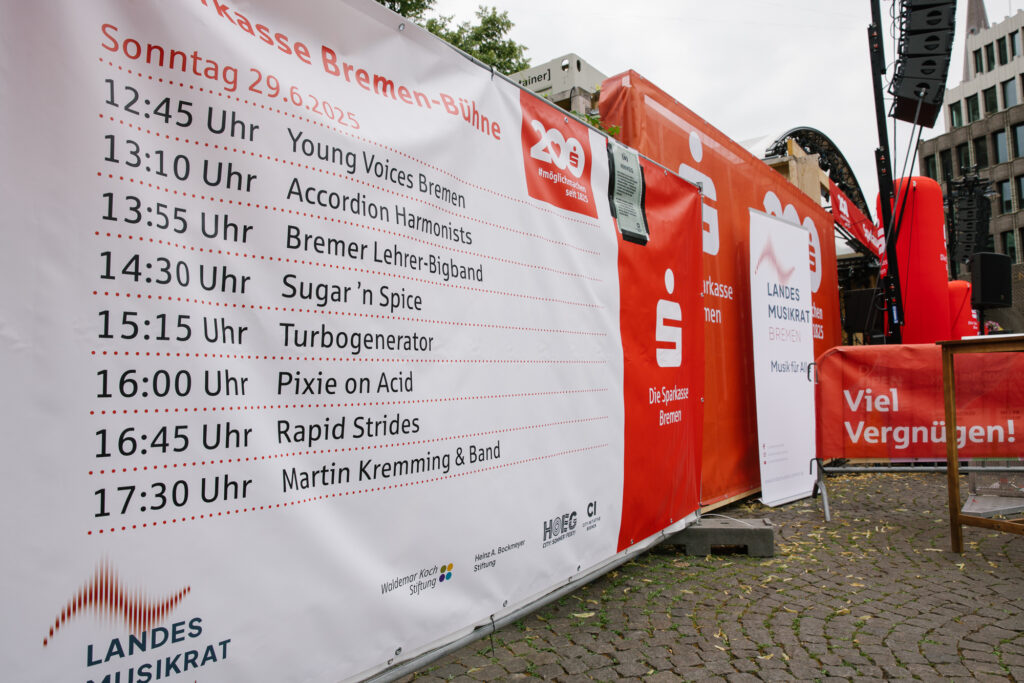 Auf einem Bauzaunbanner ist den Ablaufplan der Sparkasse Bremen-Bühne am 29.6.2025 abgedruckt. Es sind viele rote Sparkassen-Schriftzügen erkennbar. Man sieht am unteren Rand des Banners verschiedene Logos. Daneben steht ein Roll-Up mit dem Logo des Landesmusikrates Bremen.