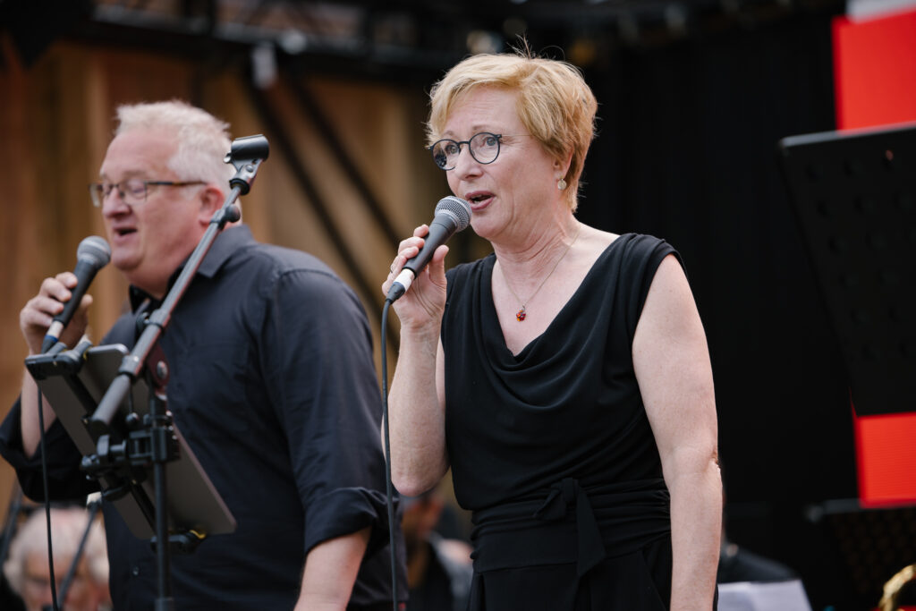 Eine Frau mit Brille, blonden kurzen Haaren und einem schwarzem Oberteil singt ins Mikrofon. Im Hintergrund ist ein weiterer Mann unscharf zu erkennen, der in ein Mikrofon singt.
