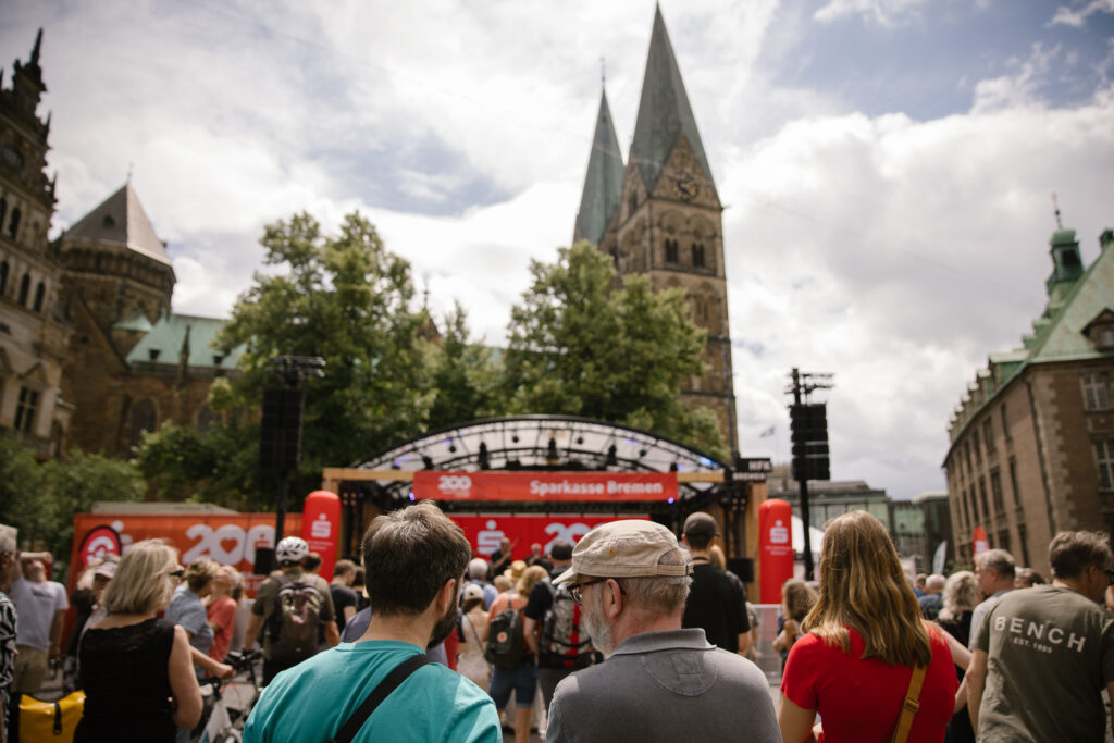 Viele Menschen stehen vor einer Open-Air Bühne, die mit roten Bannern der Sparkasse Bremen geschmückt ist. Im Hintergrund sind die Türme des Bremer Doms zu sehen.