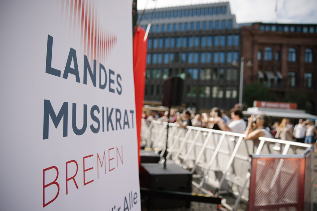 Detailaufnahme eines Banners vom landesmusikrat Bremen. Im Hintergrund ist unscharf das Publikum hinter einer Absperrung zu sehen.