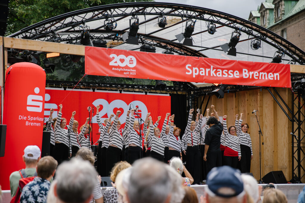 Eine Gruppe Frauen in weiß-schwarz gestreiften Oberteilen, schwarzen Hosen und roten Accessoires stehen singend neben- und hintereinander auf einer Open Air Bühne und strecken ihre Hände in die Höhe. Vor ihnen steht der schwarz gekleidete Dirigent.