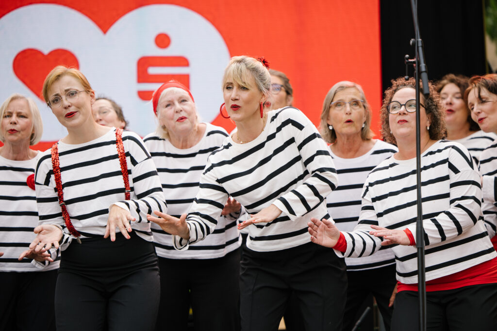 Eine Gruppe Frauen in weiß-schwarz gestreiften Oberteilen, schwarzen Hosen und roten Accessoires stehen singend nebeneinander und bewegen sich zur Musik. Im Hintergrund ist ein rotes Herz und ein rotes Sparkassenzeichen zu sehen.