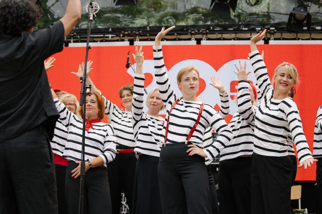 Eine Gruppe Frauen in weiß-schwarz gestreiften Oberteilen, schwarzen Hosen und roten Accessoires stehen neben- und hintereinander und strecken ihre Hände in die Höhe.