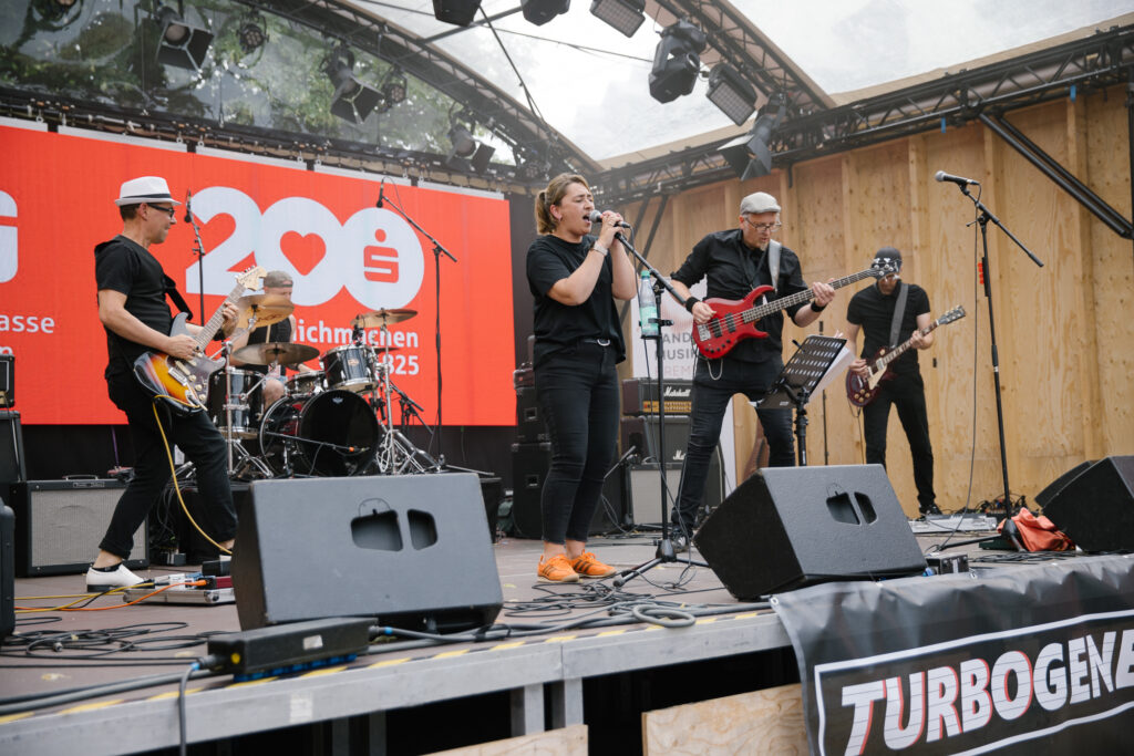 Vier Männer und eine Frau stehen singend und ihre Instrumenten spielend auf einer Open Air Bühne stehen, die mit roten Sparkassen-Schriftzügen geschmückt ist. Vor der Bühne ist ein Banner mit dem Namen der Band zu sehen.