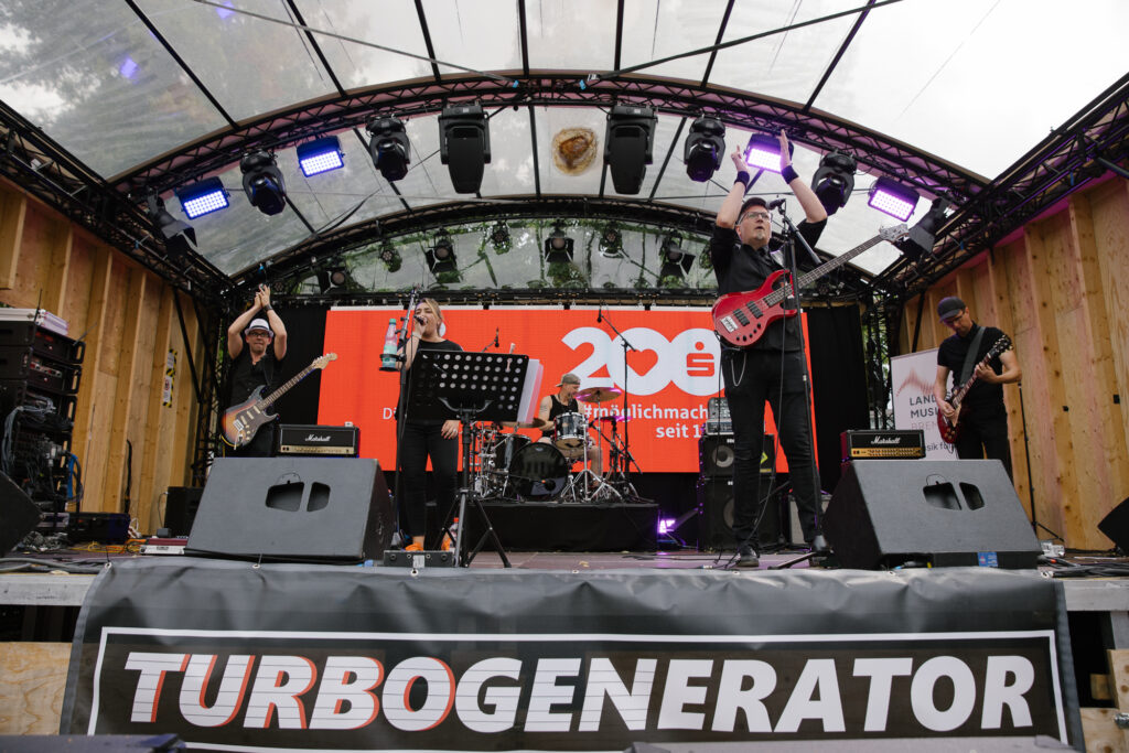 Totalaufnahme von vier Männern und einer Frau, die singend und ihre Instrumenten spielend auf einer Open Air Bühne stehen, die mit roten Sparkassen-Schriftzügen geschmückt ist. Vor der Bühne ist ein Banner mit der Aufschrift "Turbogenerator" zu sehen.