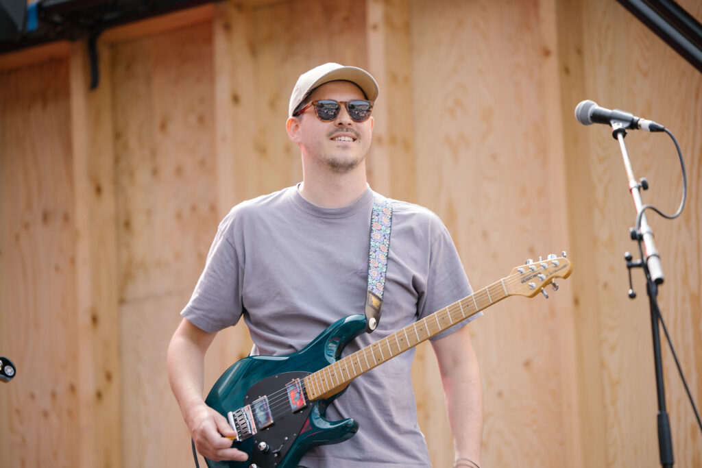 Ein Mannes mit hellem Cappie, Sonnenbrille und grauem T-Shirt steht mit seiner E-Gitarre lächelnd vor einem Mikrofon.