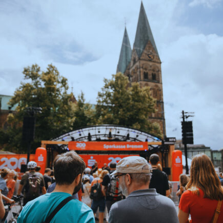 Viele Menschen stehen vor einer Open-Air Bühne, die mit roten Bannern der Sparkasse Bremen geschmückt ist. Im Hintergrund sind die Türme des Bremer Doms zu sehen.