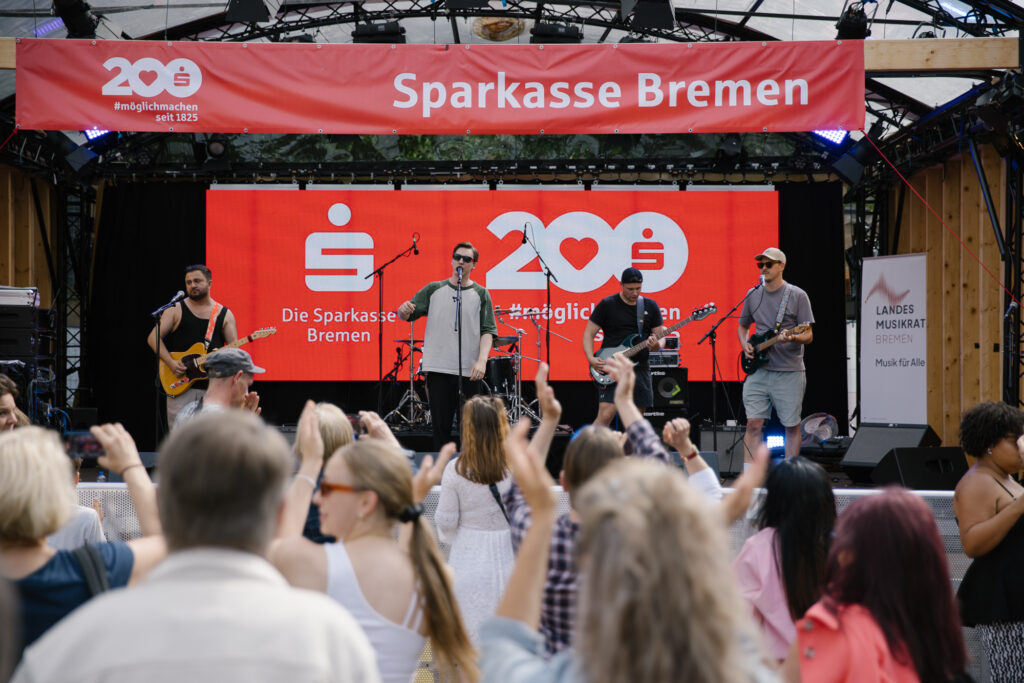 Vier Männer spielen an ihren Instrumenten auf einer Open Air Bühne, die mit roten Sparkassen-Schriftzügen geschmückt ist. Ein Mann singt ins Mikrofon. Vor der Bühne ist unscharf das Publikum zu erkennen.