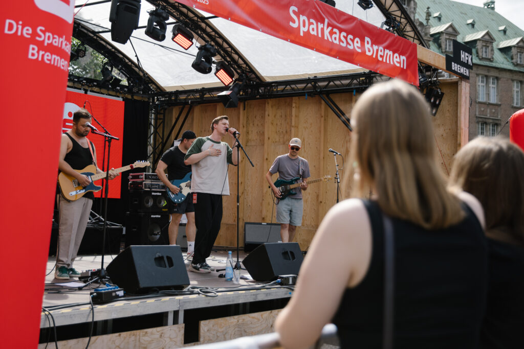 Vier Männer spielen an ihren Instrumenten auf einer Open Air Bühne, die mit roten Sparkassen-Schriftzügen geschmückt ist. Ein Mann singt ins Mikrofon.