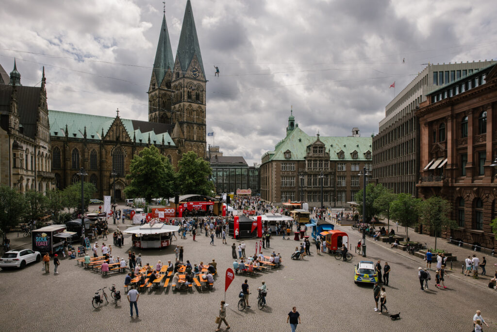 Totalaufnahme des Domshofes in Bremen. Menschen stehen vor einer Open-Air Bühne, die mit roten Bannern der Sparkasse Bremen geschmückt ist. Auf Bierbänken und an Gastronomie-Ständen sind viele weitere Personen zu sehen. Im Hintergrund ist der Bremer Dom und weitere Gebäude zu erkennen. In luftiger Höhe balancieren zwei Menschen auf Slacklines.