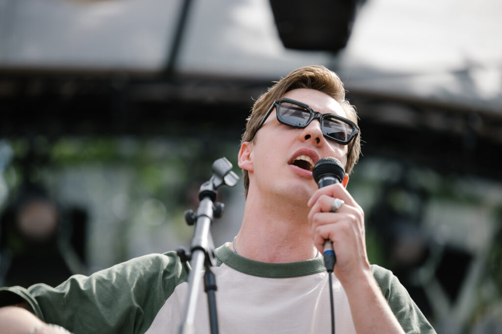 Detailaufnahme eines Mannes mit kurzen hellbraunen Haaren, Sonnenbrille und grün-weißem T-Shirt, der leidenschaftlich ins Mikrofon singt.