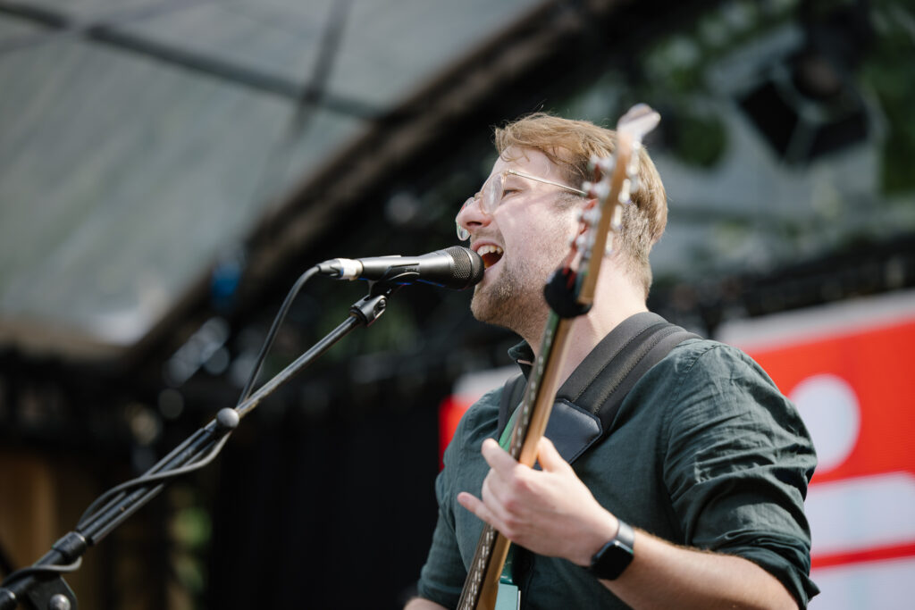 Ein Mann mit kurzen hellbraunen Haaren, Brille und in dunklem Hemd singt leidenschaftlich ins Mikrofon.